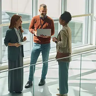 Three colleagues standing together in an office building, one holding an open laptop.