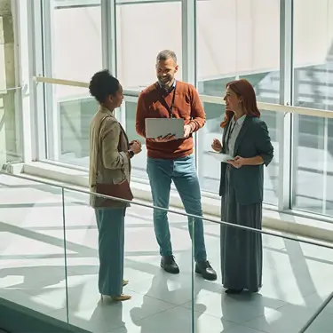 Three colleagues standing together in an office building, one holding an open laptop.