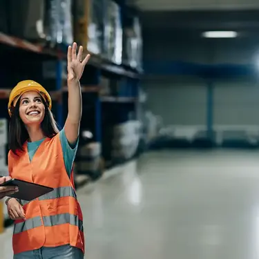 Two smiling colleagues in reflective vests and helmets reviewing warehouse stock using a tablet.